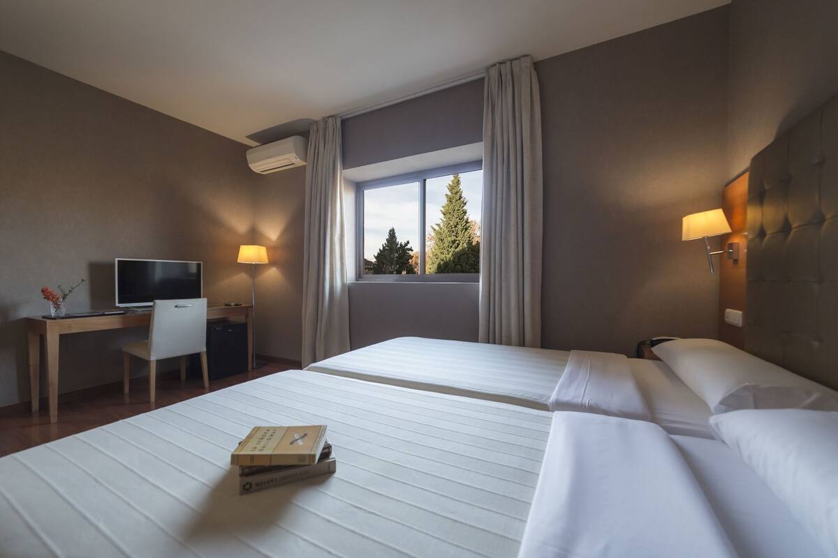 Habitación de hotel con dos camas, un escritorio con computadora, televisor y ventana con vista a árboles.