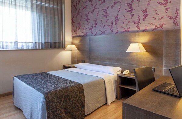 Una habitación de hotel con una cama, dos lámparas de noche, un escritorio con una computadora portátil y un teléfono.