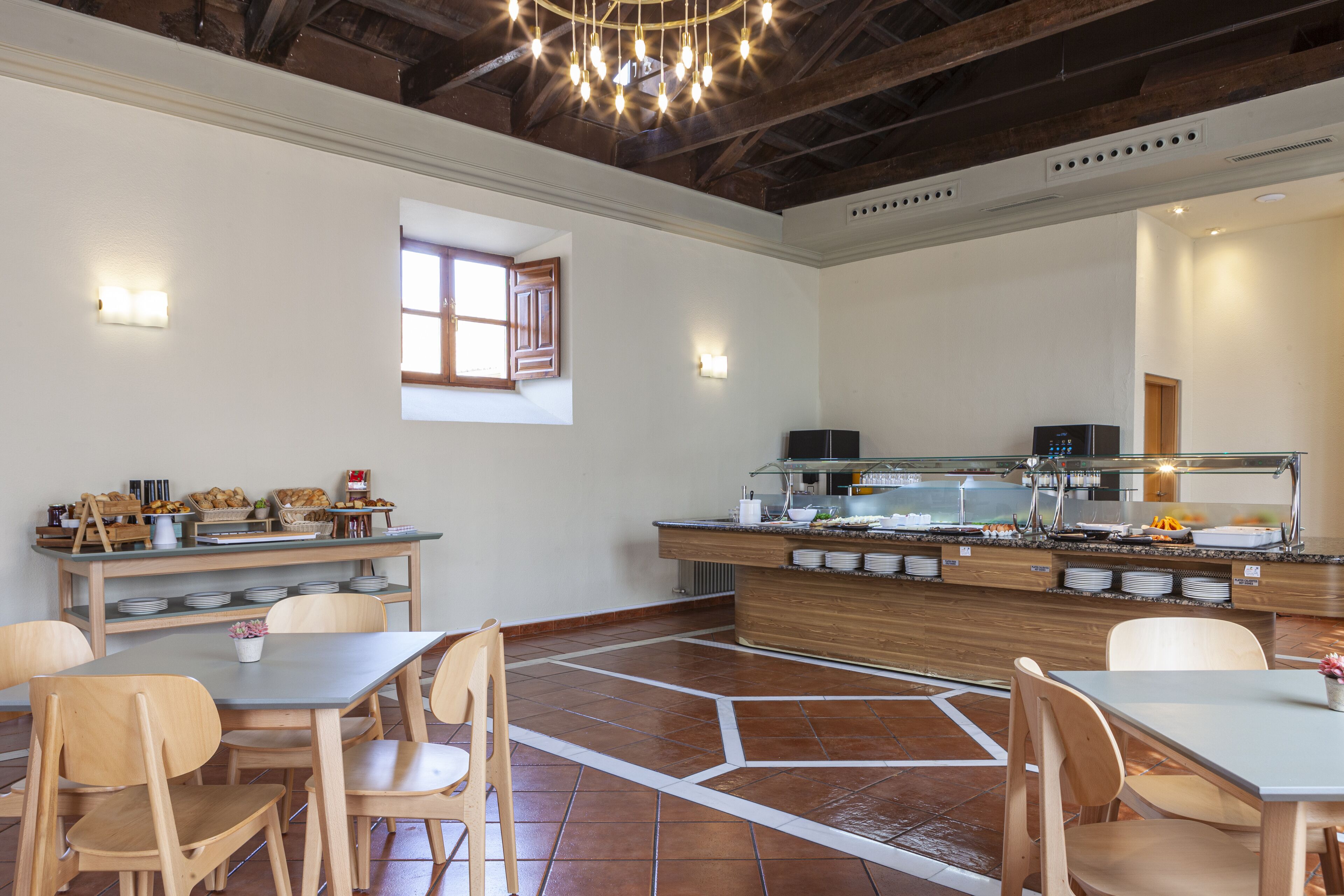 A dining area with wooden tables and chairs, a buffet counter with food items, and a window with wooden shutters.
