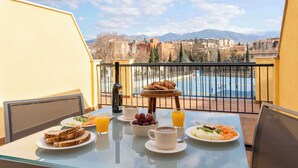 A balcony with breakfast setup including toast, fruit, and coffee, overlooking a pool and mountains.