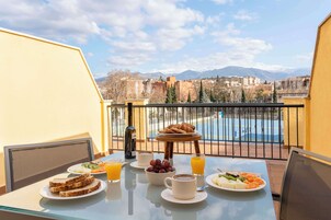 A balcony with breakfast setup including toast, fruit, and coffee, overlooking a pool and mountains.