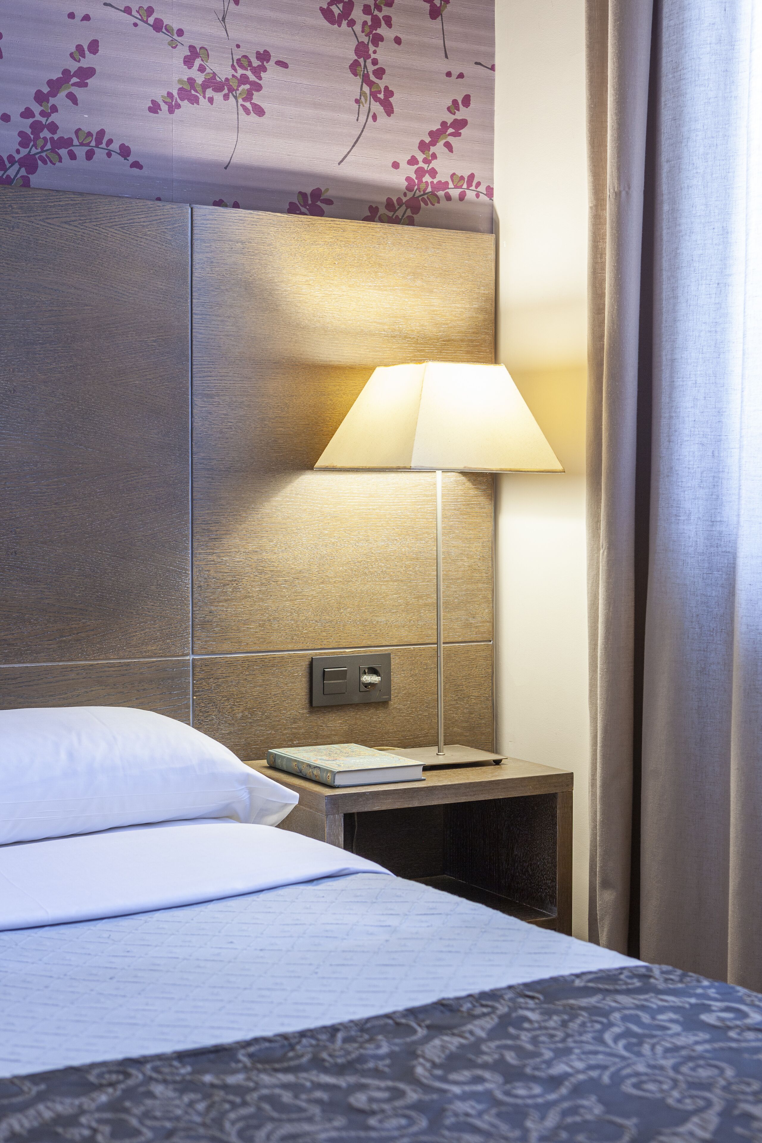 Une chambre d’hôtel avec un lit, une table de chevet avec une lampe, un livre et un mur orné d’un motif floral.