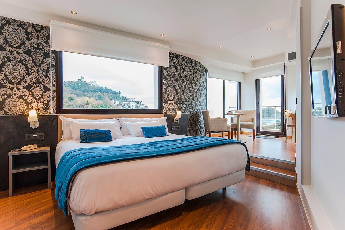 Un dormitorio moderno con una cama grande, una mesita de noche, un escritorio y una silla. Hay un ventanal con vistas a un paisaje urbano.