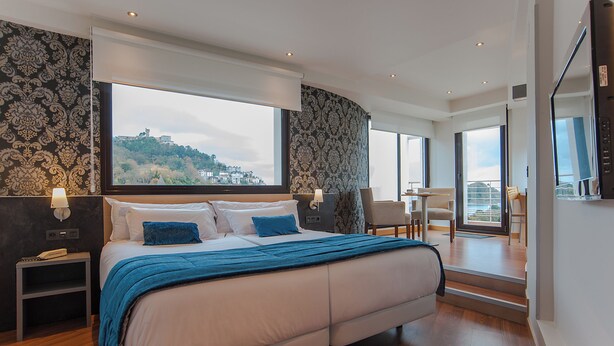 A modern bedroom with a large bed, a nightstand, a chair, and a view of a cityscape through large windows. - Sercotel Codina (San Sebastián)