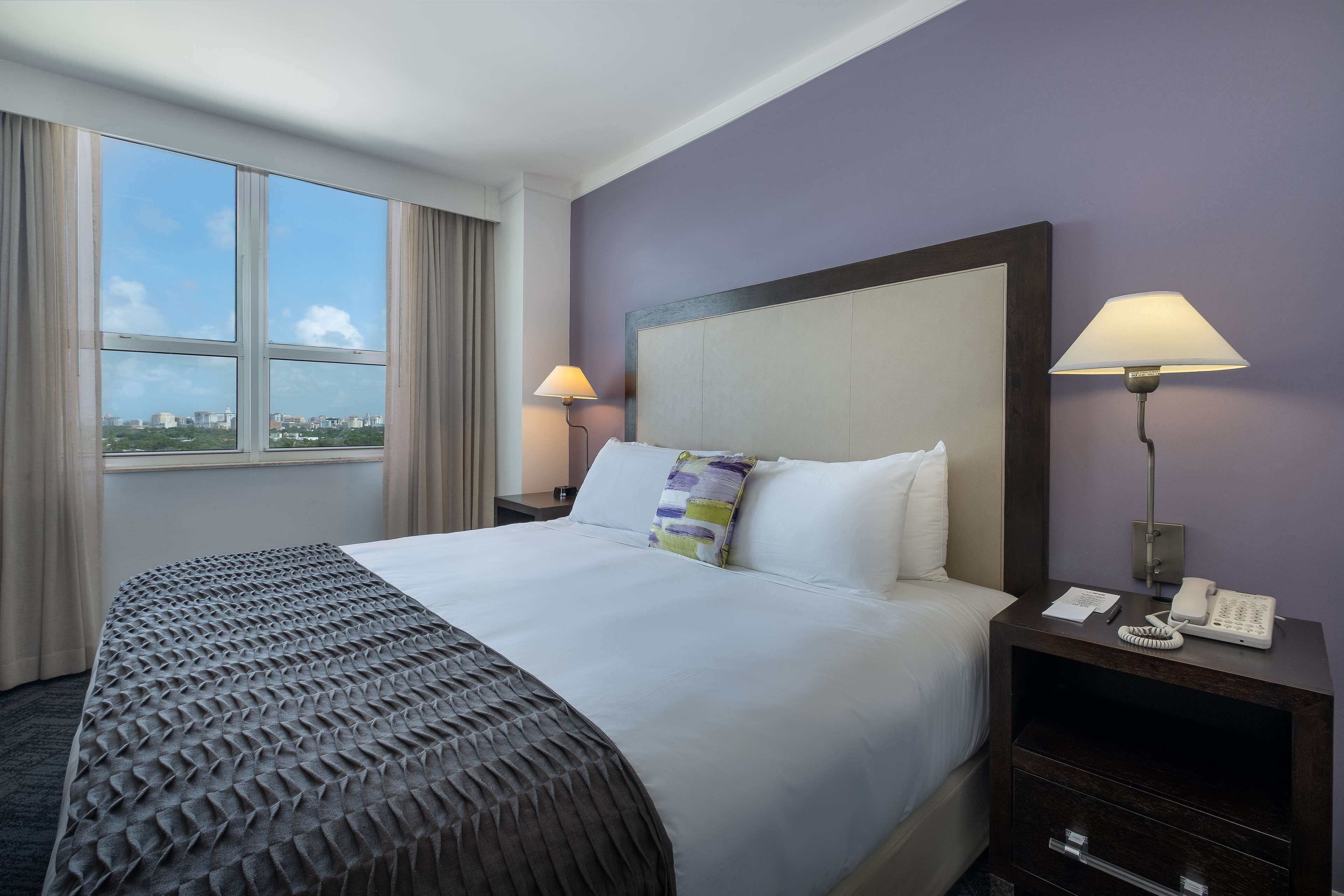 A hotel room with a large bed, two bedside lamps, a nightstand with a phone, and a window with curtains.