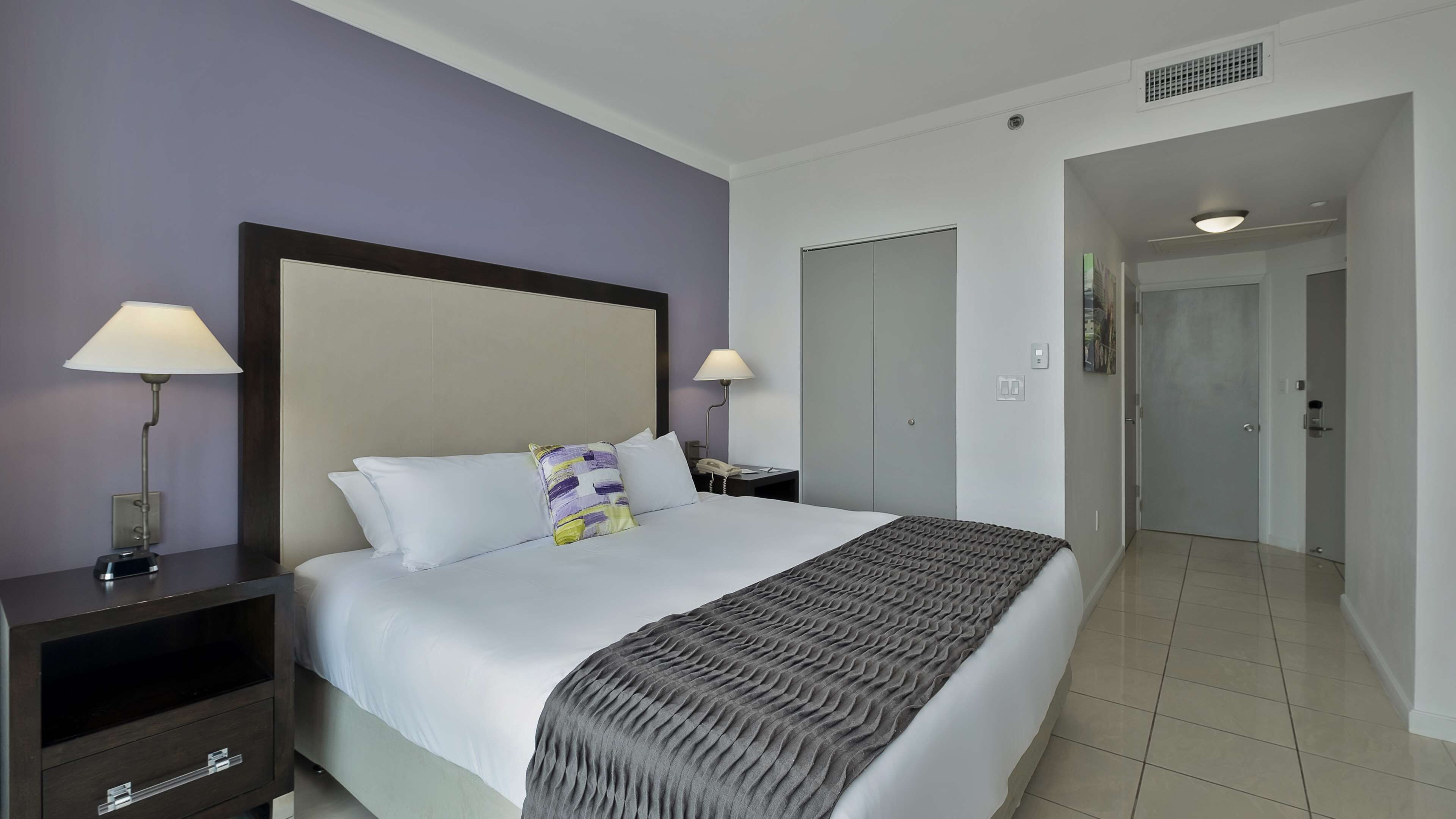 A hotel room with a bed, two bedside lamps, a nightstand, and a door leading to another room.