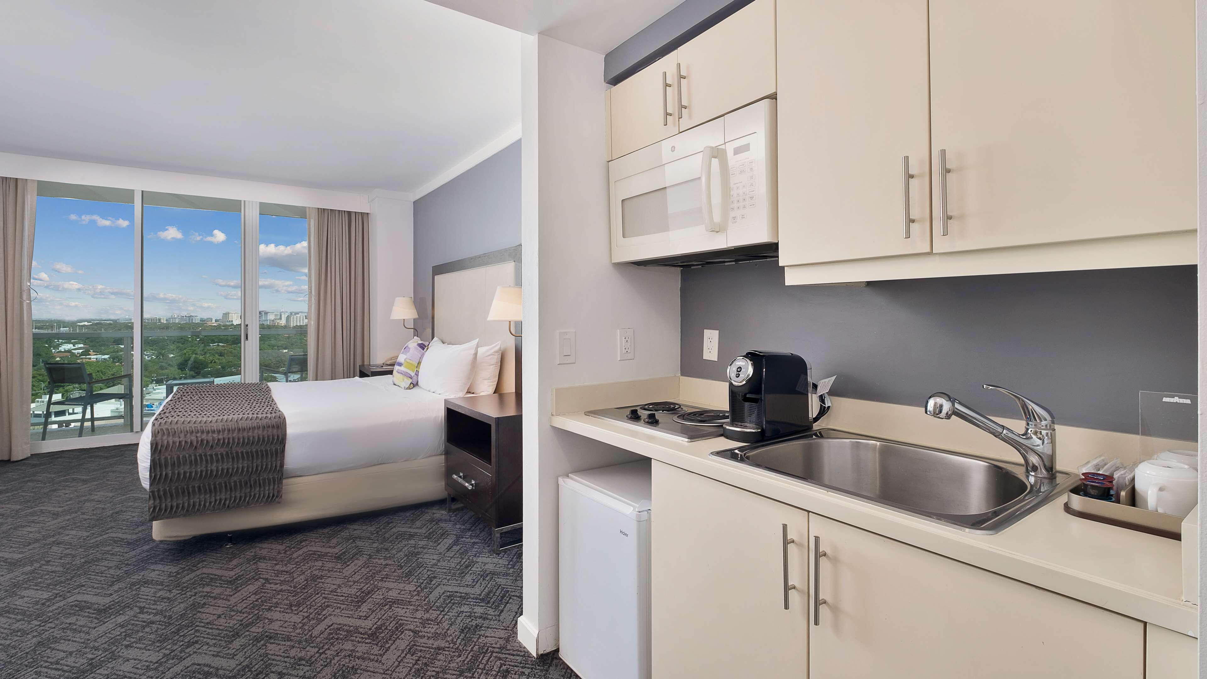 A hotel room with a bed, a small kitchenette, a view of the city, and a window with curtains.
