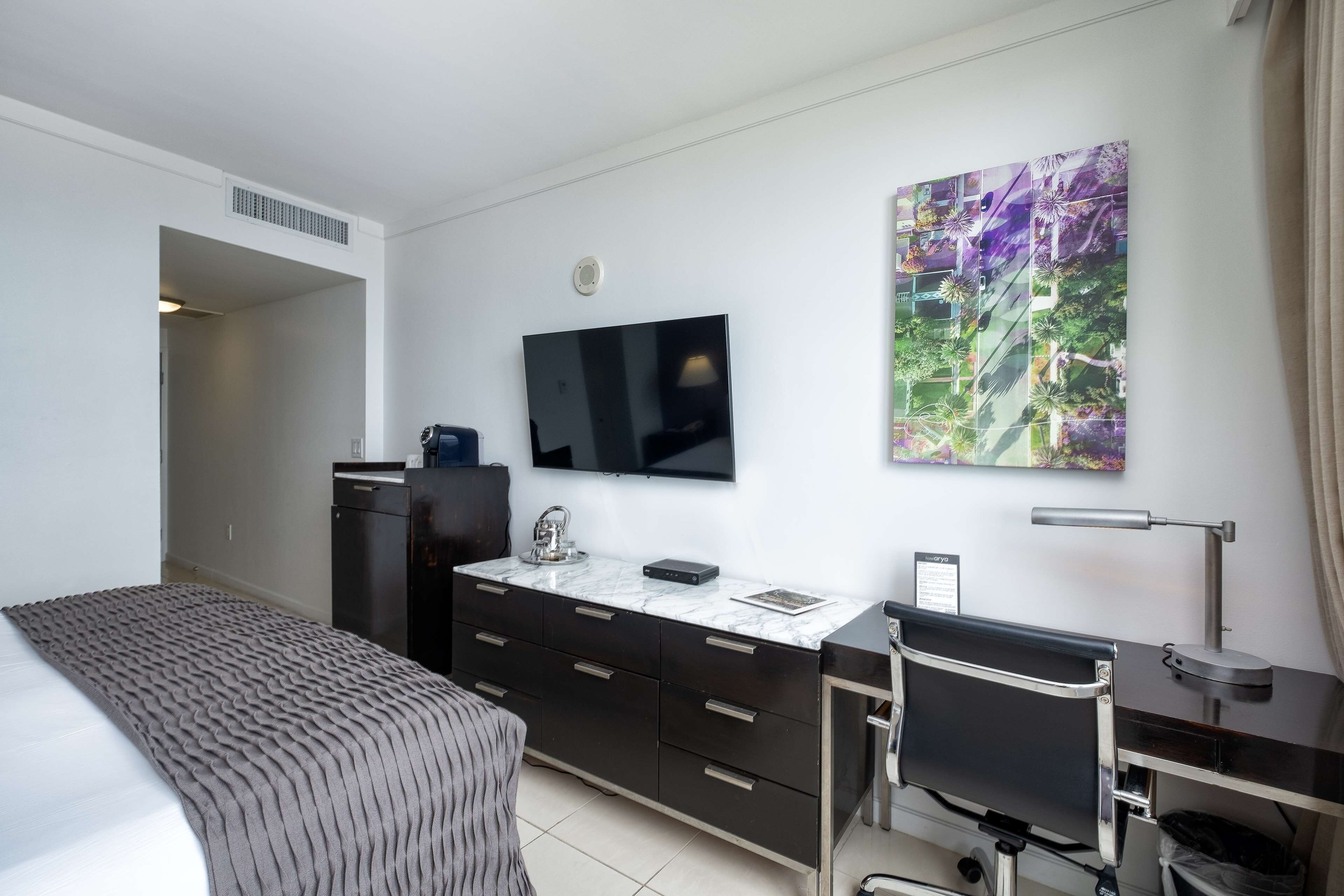 Ein Hotelzimmer mit einem Bett, einem Schreibtisch mit Stuhl, einem Fernseher, einer Kommode mit Marmorplatte und einem großen abstrakten Gemälde an der Wand.
