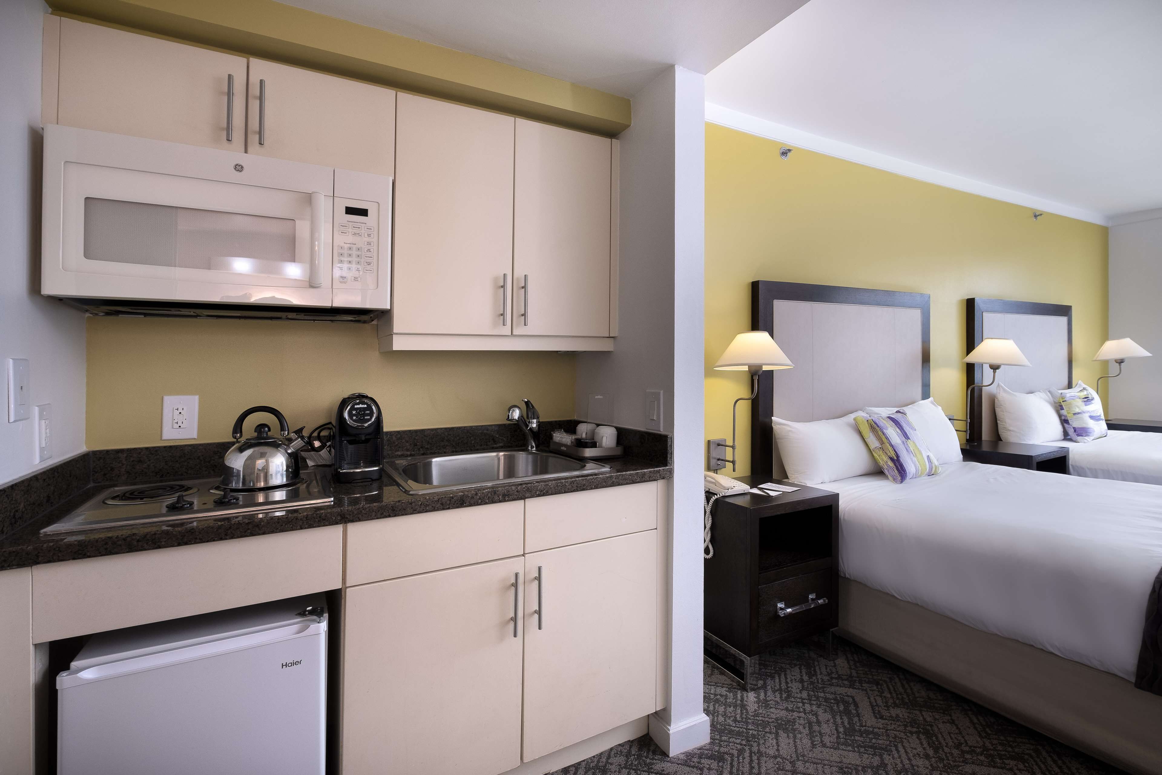 A hotel room with a kitchenette, including a microwave, stove, and refrigerator. There are two beds with nightstands, each with a lamp and a phone.