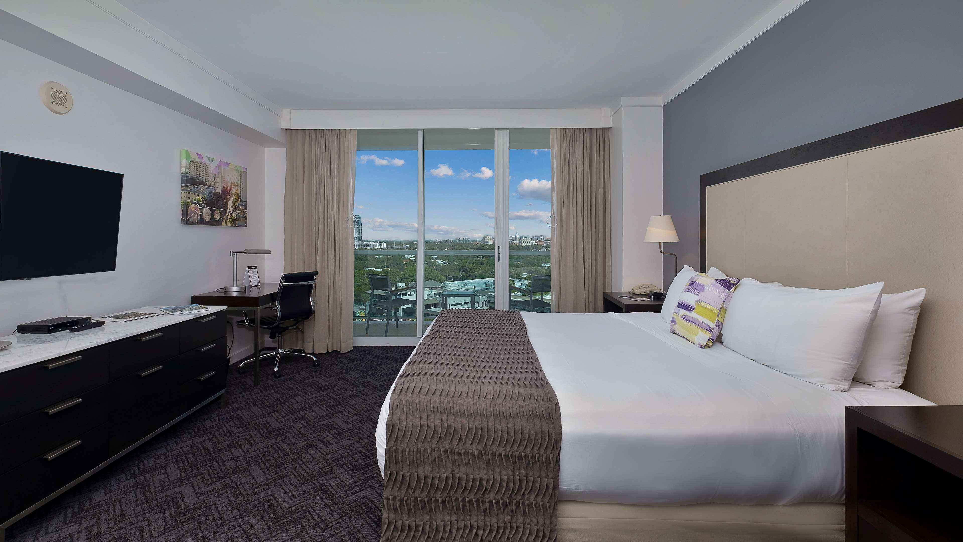 A hotel room with a large bed, a desk with a computer, a TV, and a view of the city through large windows.