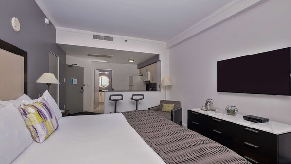 A hotel room with a bed, a desk, a chair, a TV, and a kitchen area in the background.