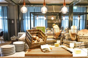 A breakfast buffet setup with various breads and pastries in baskets, plates stacked on the counter, and hanging light bulbs.
