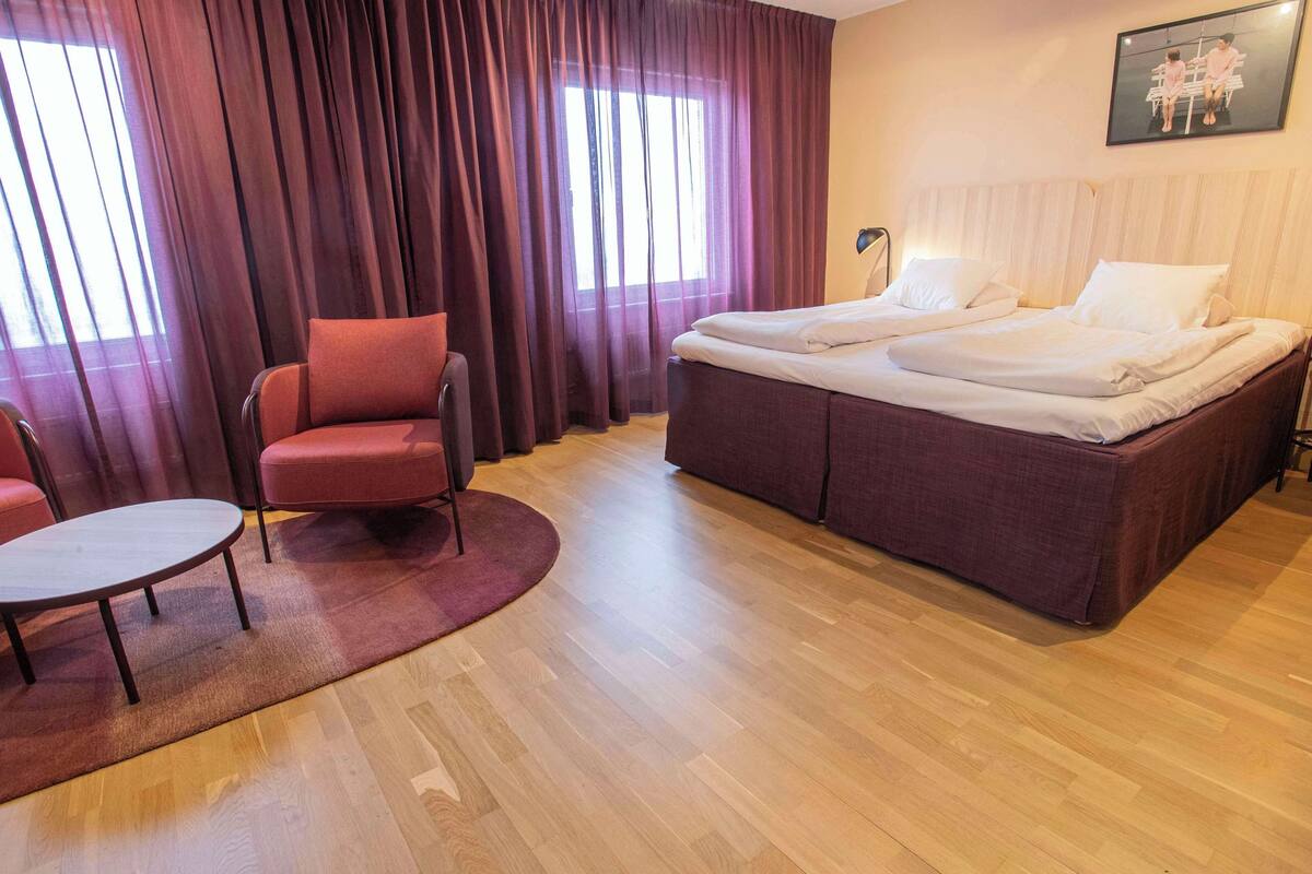 Ein Hotelzimmer mit Bett, Sessel, kleinem Tisch und einem Fenster mit Vorhängen.