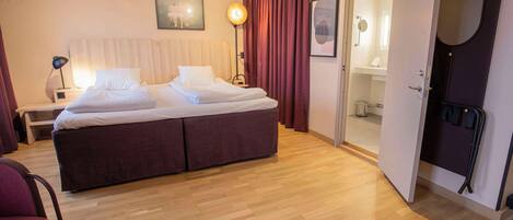 A hotel room with a bed, a nightstand, a lamp, a chair, and a door leading to a bathroom.