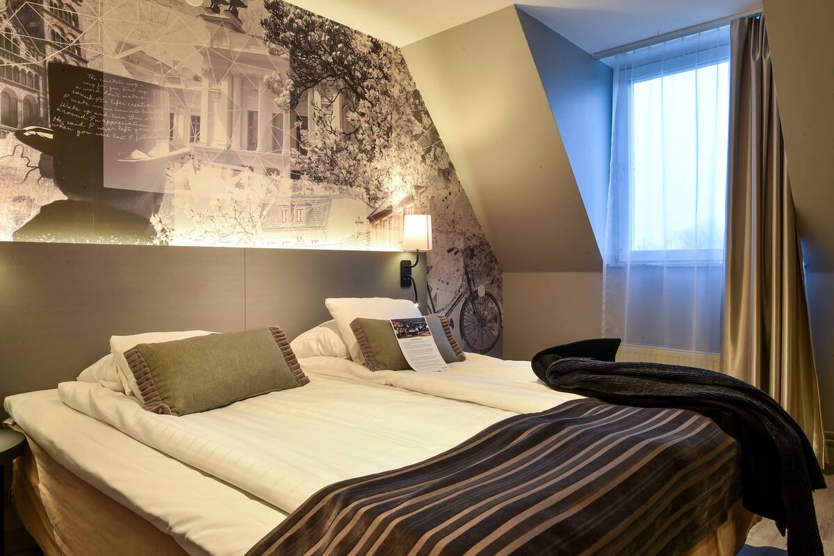 Ein Hotelzimmer mit einem großen Bett, einem Kopfteil mit Stadtbild-Wandgemälde, einem Fenster mit Vorhängen und einer Lampe.