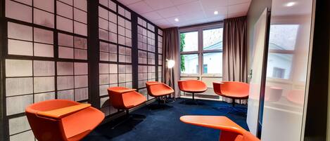 A conference room with a blue carpet, orange chairs, a large window with curtains, and a view of the outside.