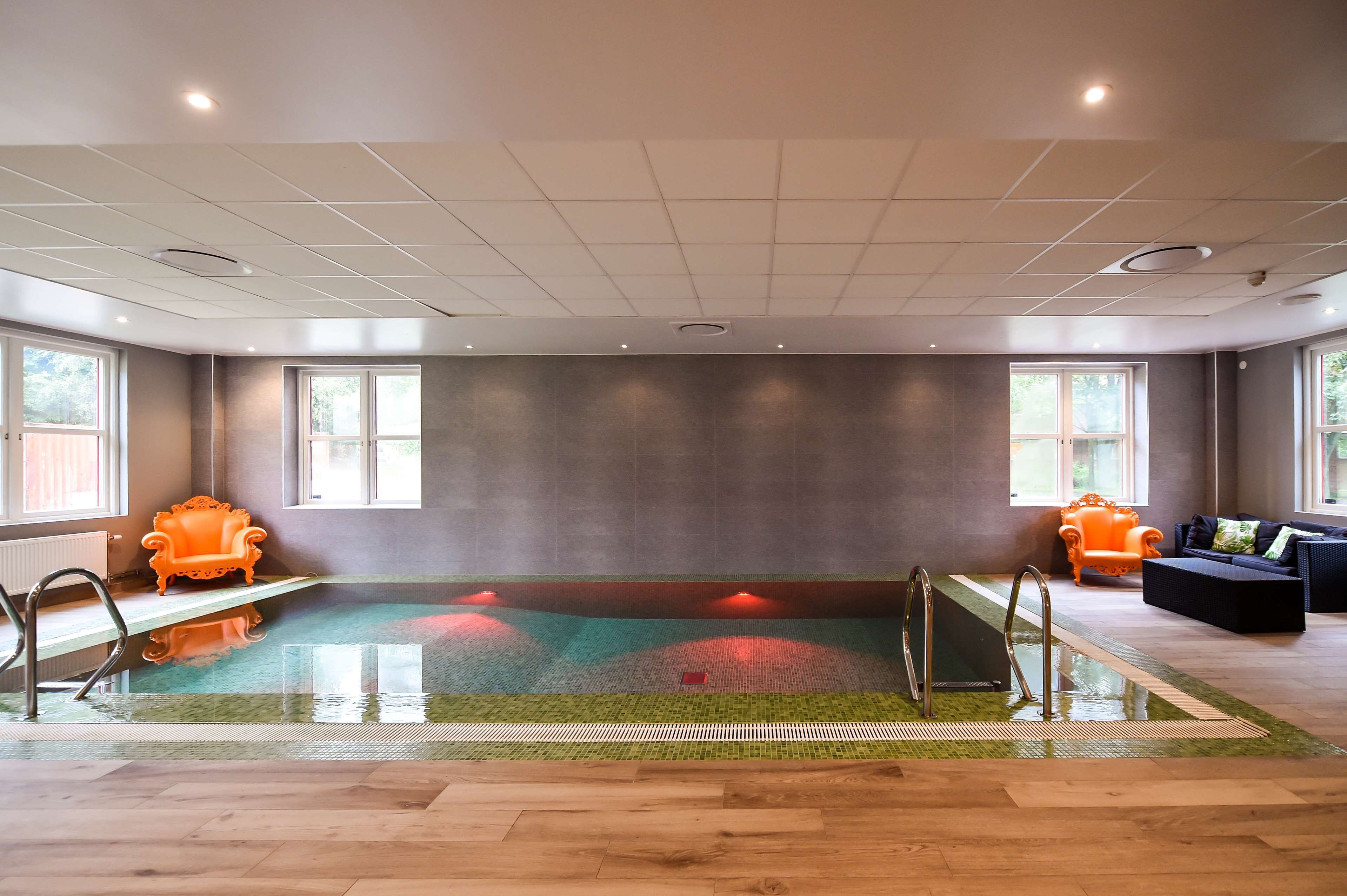 A modern indoor swimming pool with a glass floor, surrounded by wooden flooring and furnished with two orange armchairs.