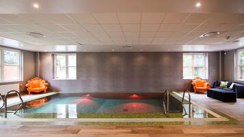 A modern indoor swimming pool with a glass floor, surrounded by wooden flooring and furnished with two orange armchairs.
