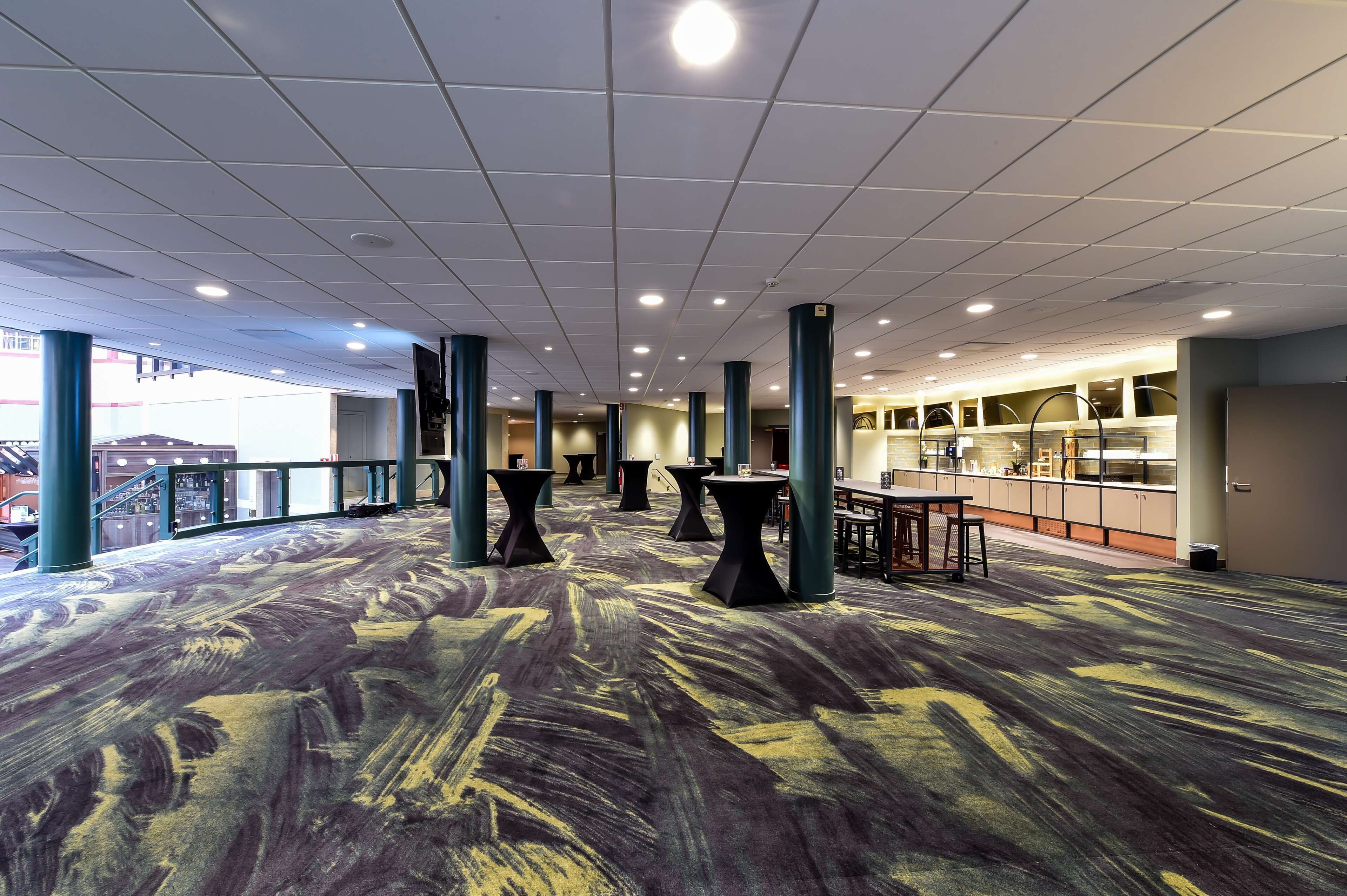 A spacious indoor area with a patterned carpet, multiple tables and chairs, and a kitchen area in the background.