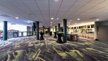 A spacious indoor area with a patterned carpet, multiple tables and chairs, and a kitchen area in the background.