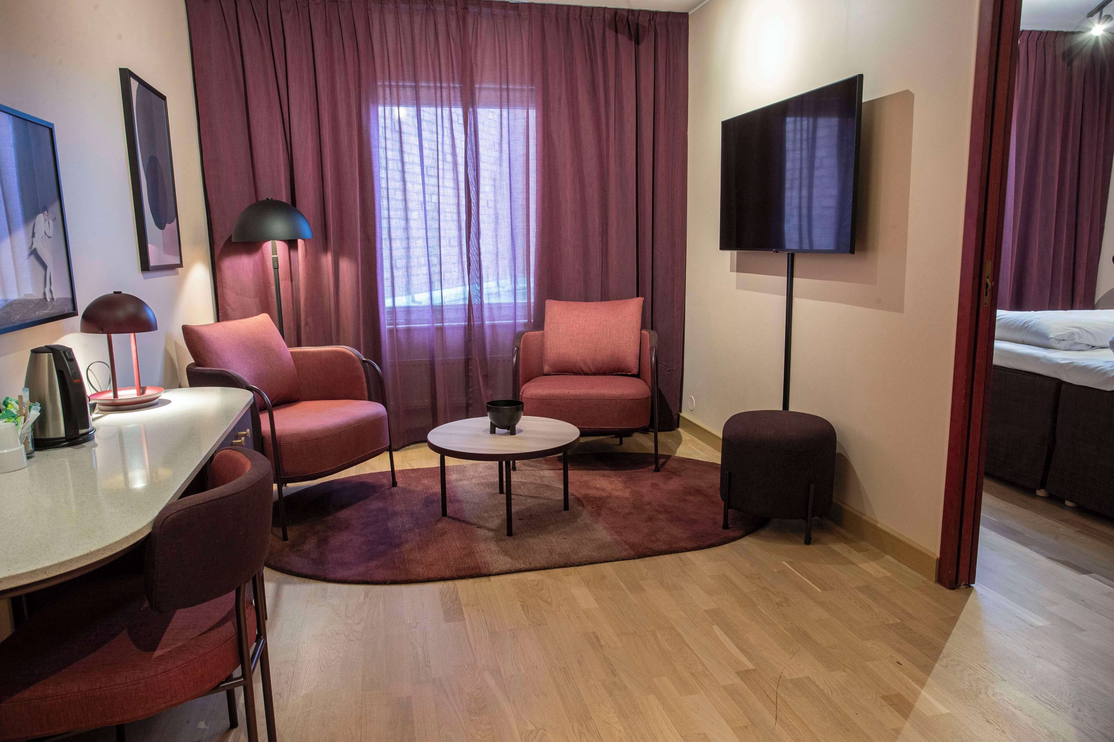 Habitación de hotel con una cama, dos sillones, una mesita de café, un televisor y un escritorio con una lámpara.