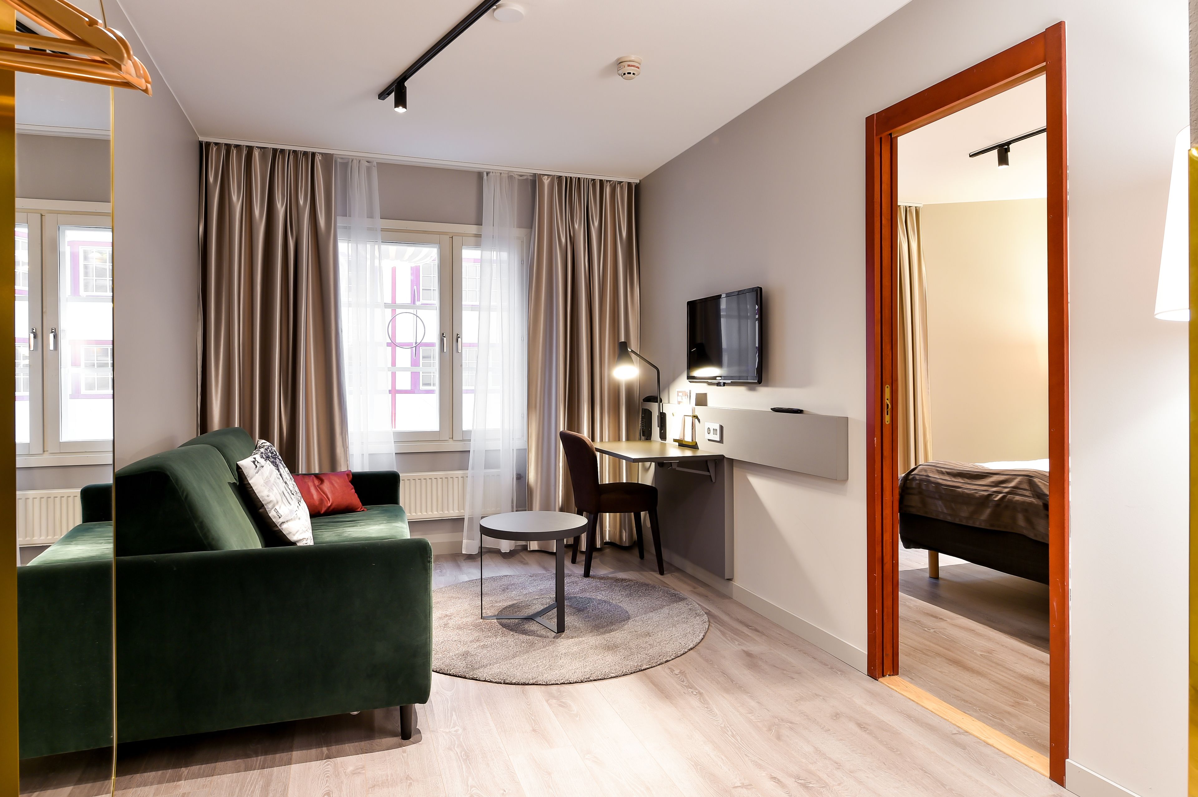 Una habitación de hotel moderna con un sofá verde, una mesa redonda, un escritorio con una silla, una cama y un televisor montado en la pared.