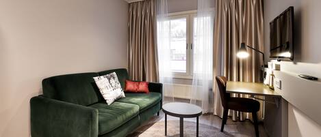A modern living room with a green sofa, a small round table, a desk with a chair, and a TV mounted on the wall.