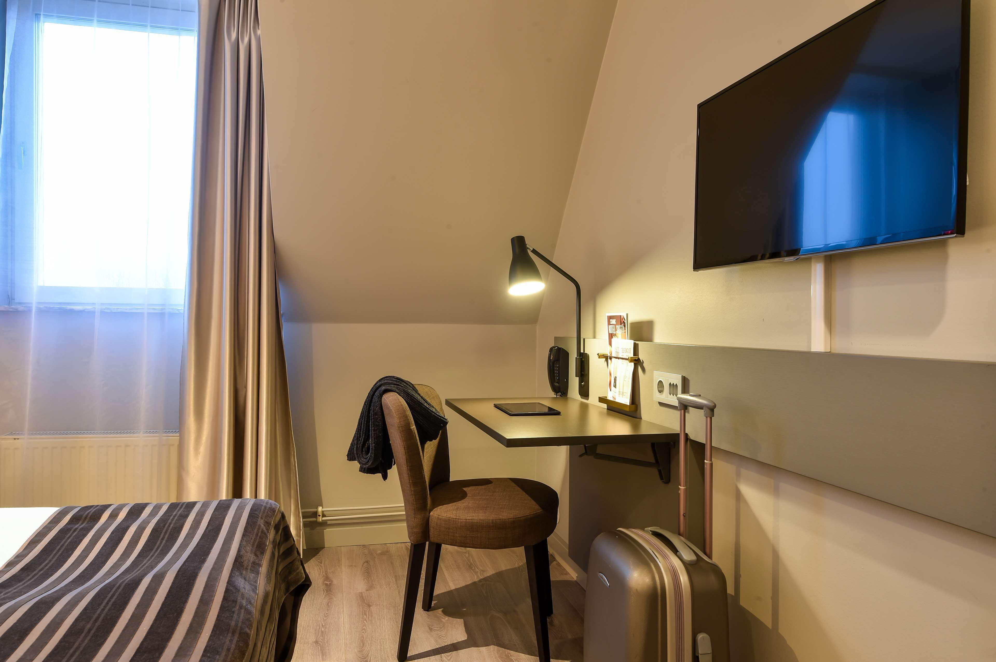 A hotel room with a bed, a desk with a chair, a television, a lamp, a suitcase, and a window with curtains.