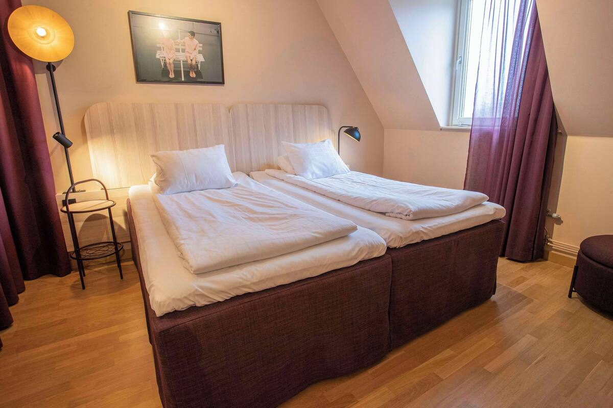 Ein Schlafzimmer mit einem großen Bett, einem Nachttisch, einer Stehlampe und einem Fenster mit Vorhängen.