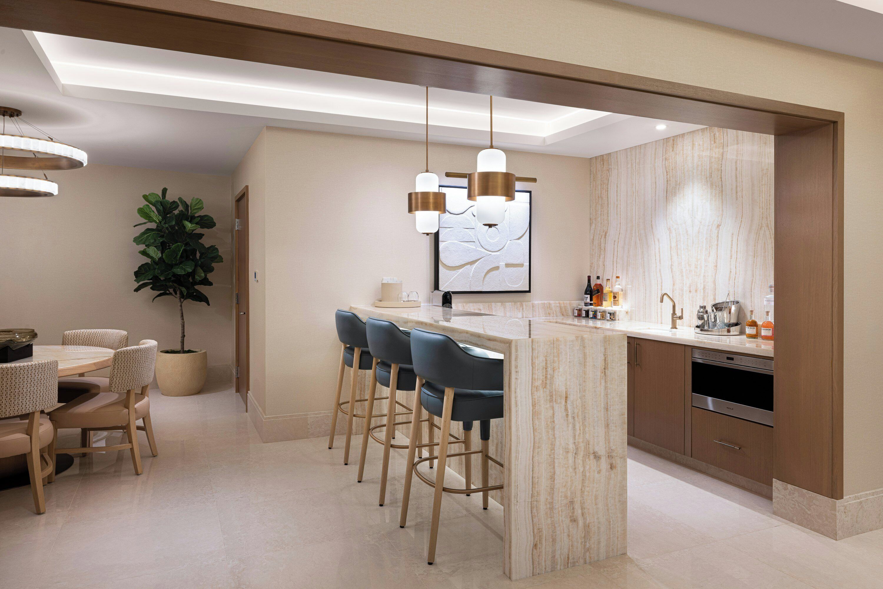 A modern kitchen with a marble countertop, bar stools, pendant lights, and a potted plant.