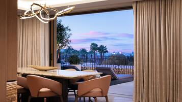 Un espace de repas moderne avec une table et des chaises, une vue sur des palmiers et le paysage urbain à travers une grande fenêtre, et un balcon avec une balustrade.