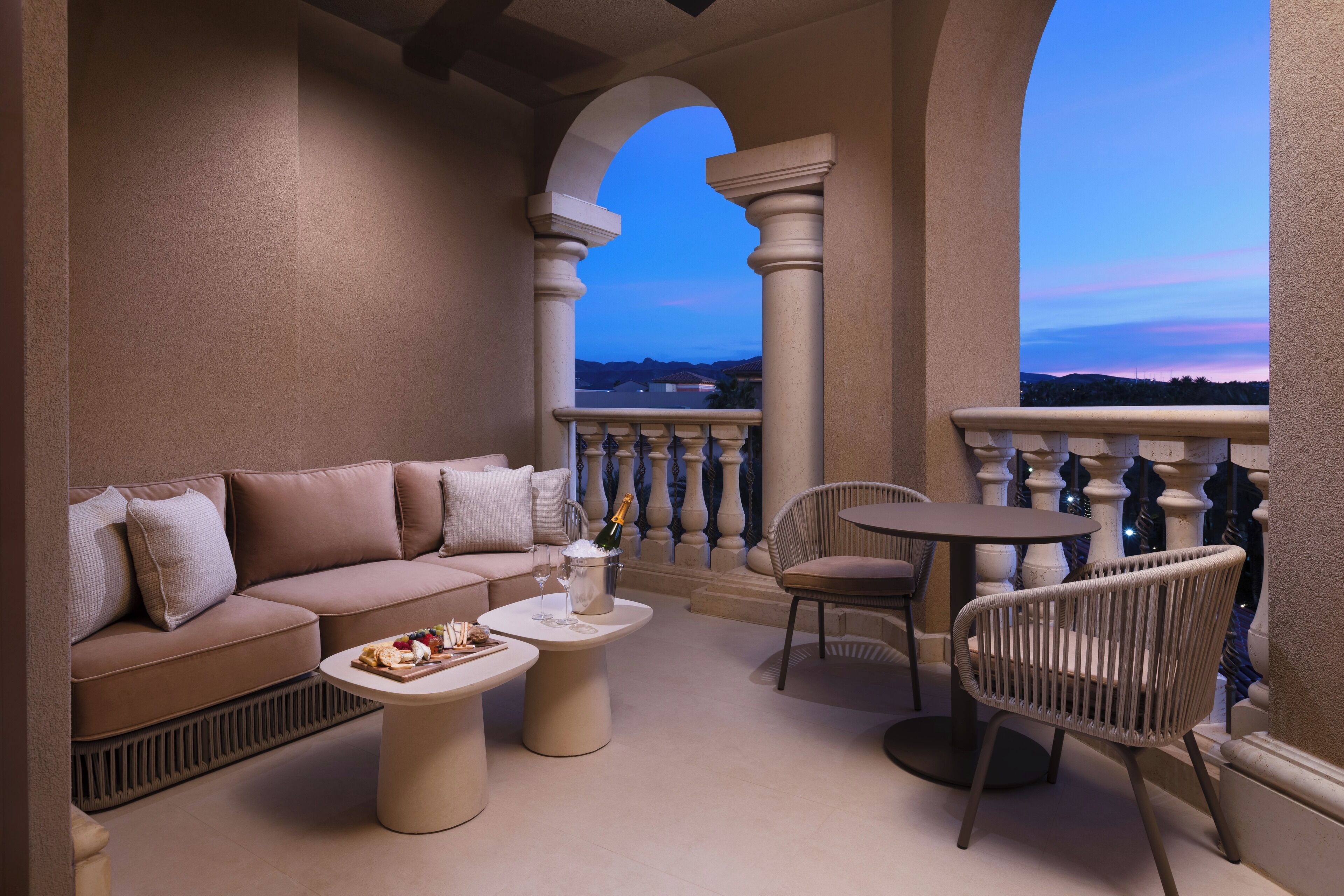 A balcony with a beige sofa, a round table, and a wicker chair, overlooking a scenic view.