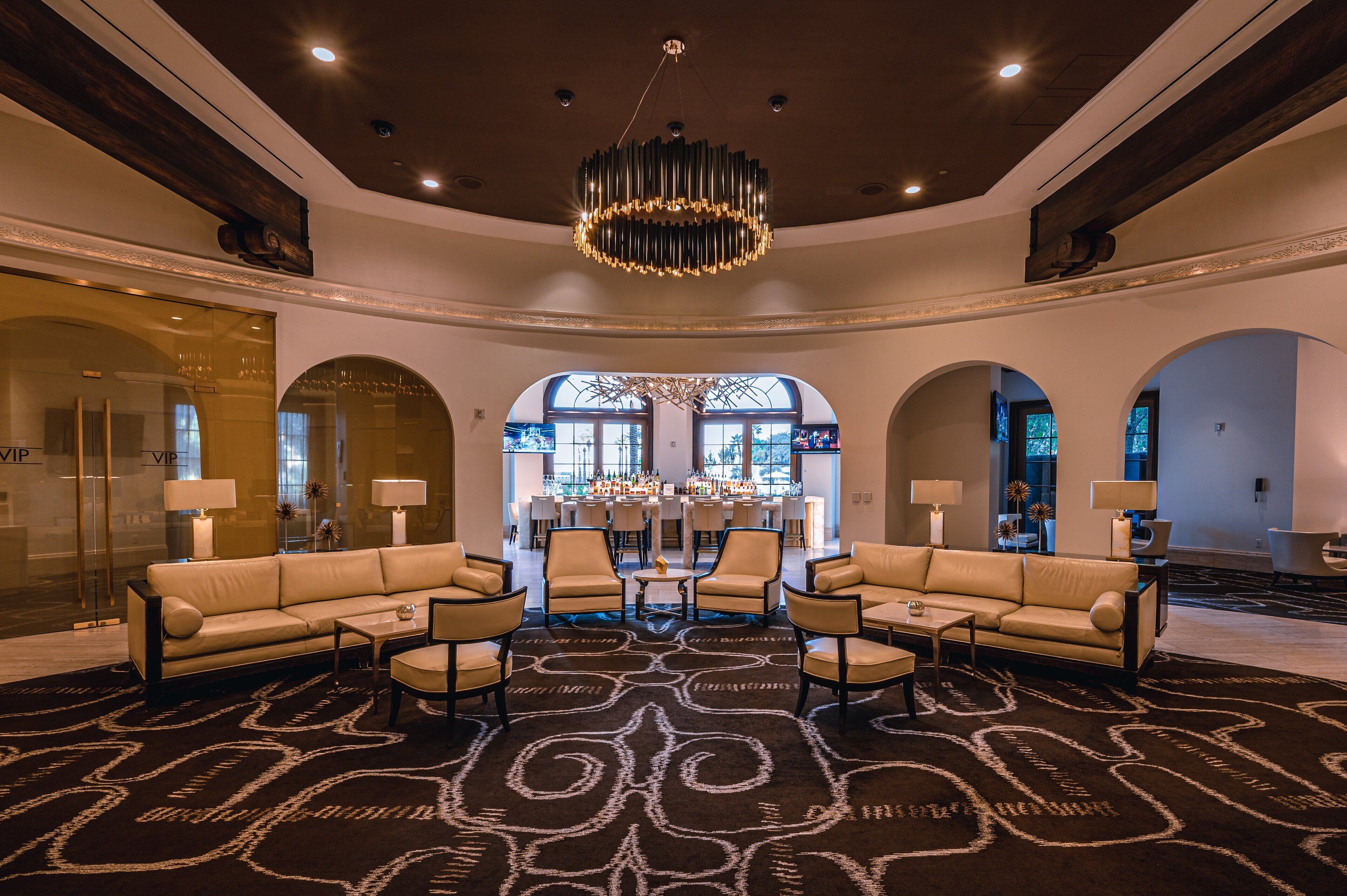 A spacious lobby with a central seating area, a bar in the background, and a large chandelier.