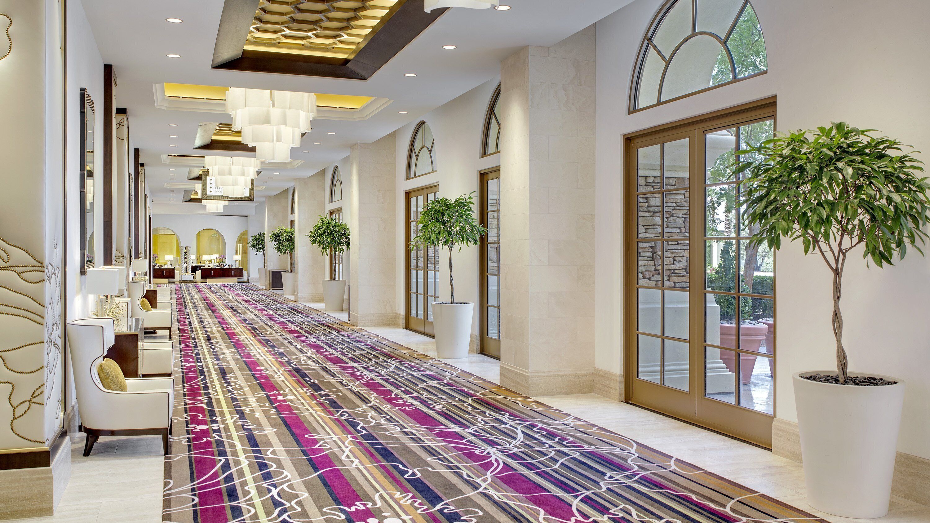 A hotel corridor with a patterned carpet, seating area, potted plants, and large windows.