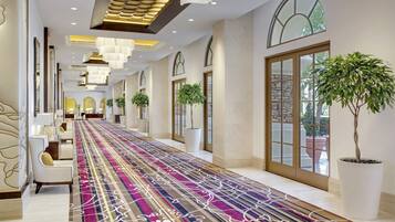 A hotel corridor with a patterned carpet, seating area, potted plants, and large windows.