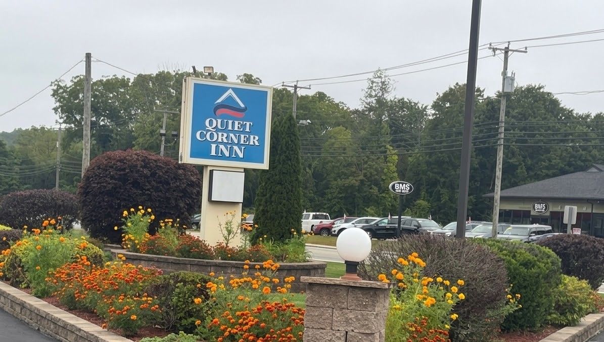 Letrero del Quiet Corner Inn con un logotipo, rodeado de macizos de flores y un muro de piedra.