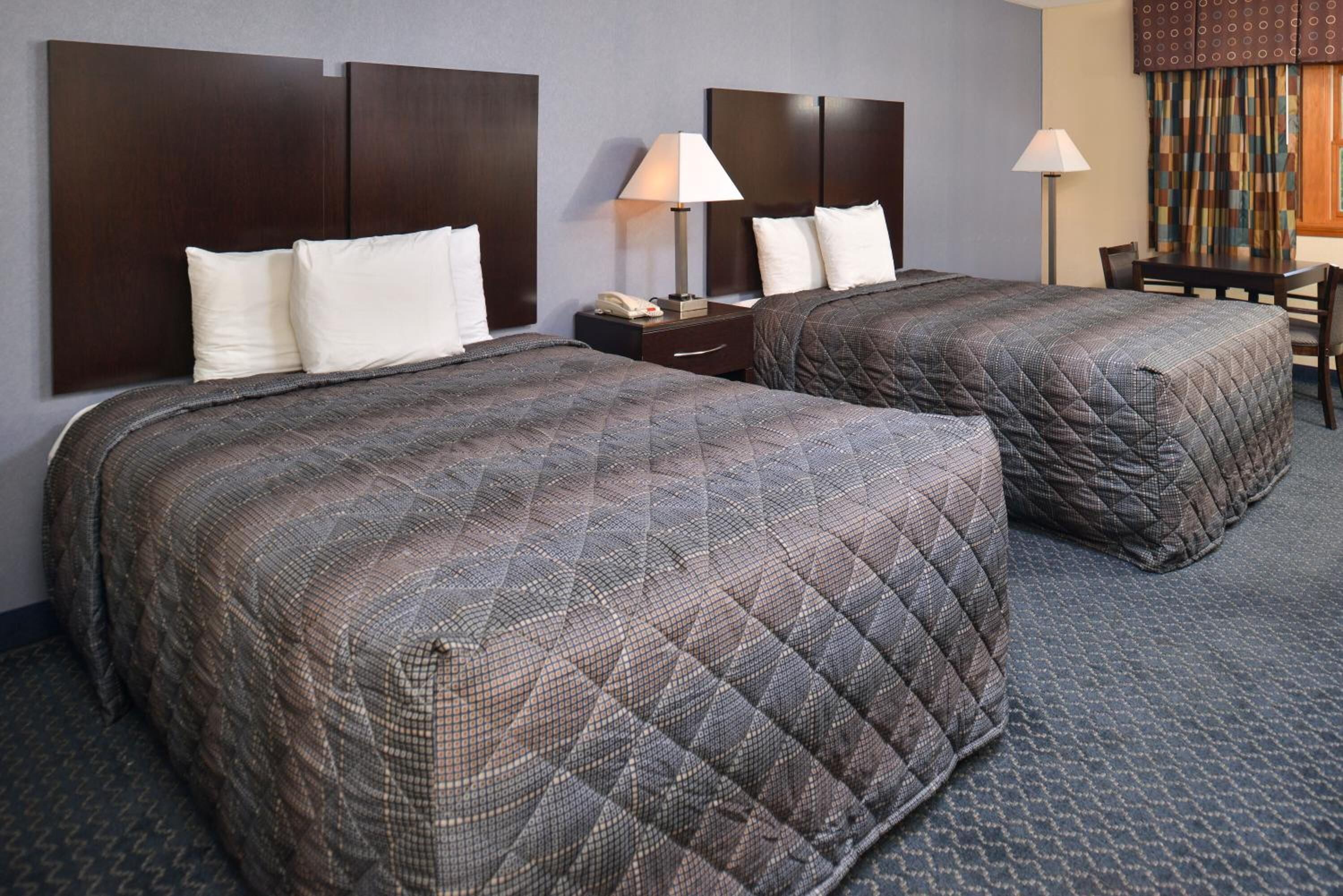 Habitación de hotel con dos camas tamaño queen, cabeceros de madera oscura, un escritorio pequeño con lámpara y una mesa de comedor con sillas.