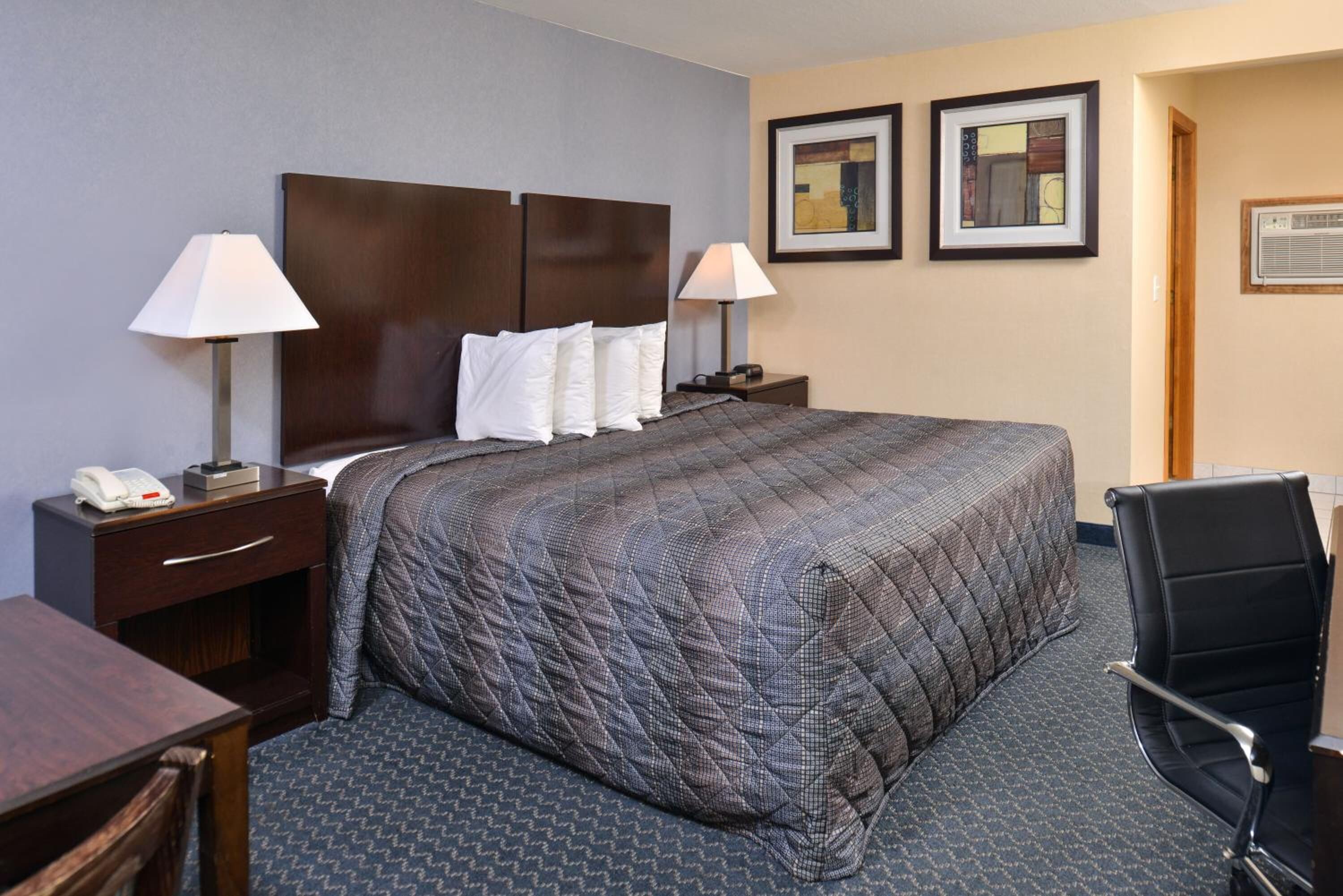 A hotel room with a large bed, two bedside tables with lamps, a desk, and a chair. There are two framed pictures on the wall.
