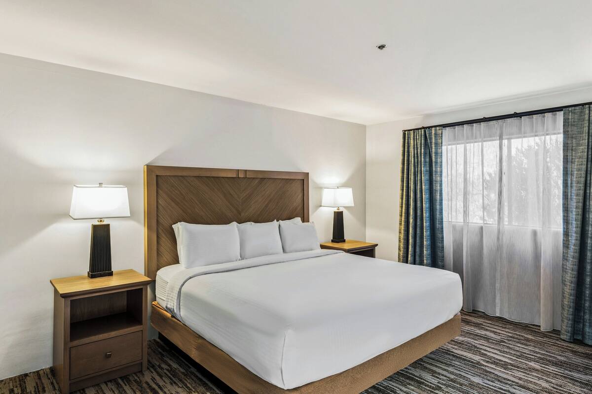 A hotel room with a bed, two bedside tables with lamps, a wooden headboard, and a large window with curtains.