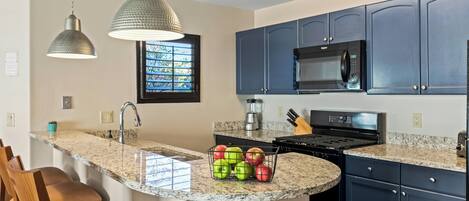 A kitchen with a granite countertop, a built-in microwave, and a stove. There are wooden chairs and a window with blinds.
