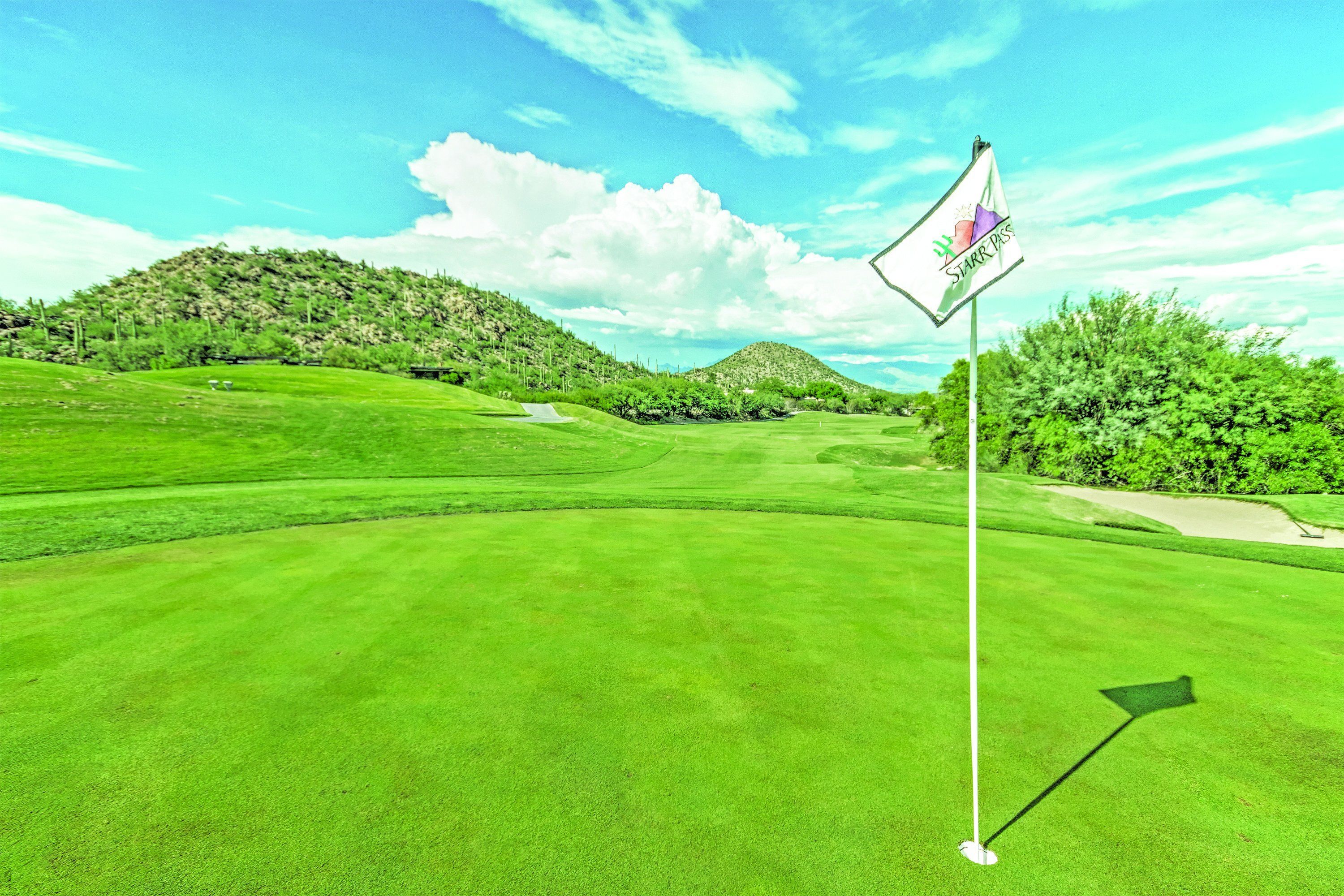 Un terrain de golf avec un drapeau sur un mât, entouré de collines verdoyantes et d’arbres.