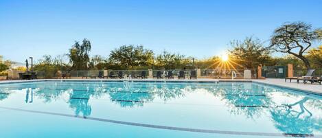 En klarblå pool med solstolar och ett staket, omgiven av träd och en klar himmel.