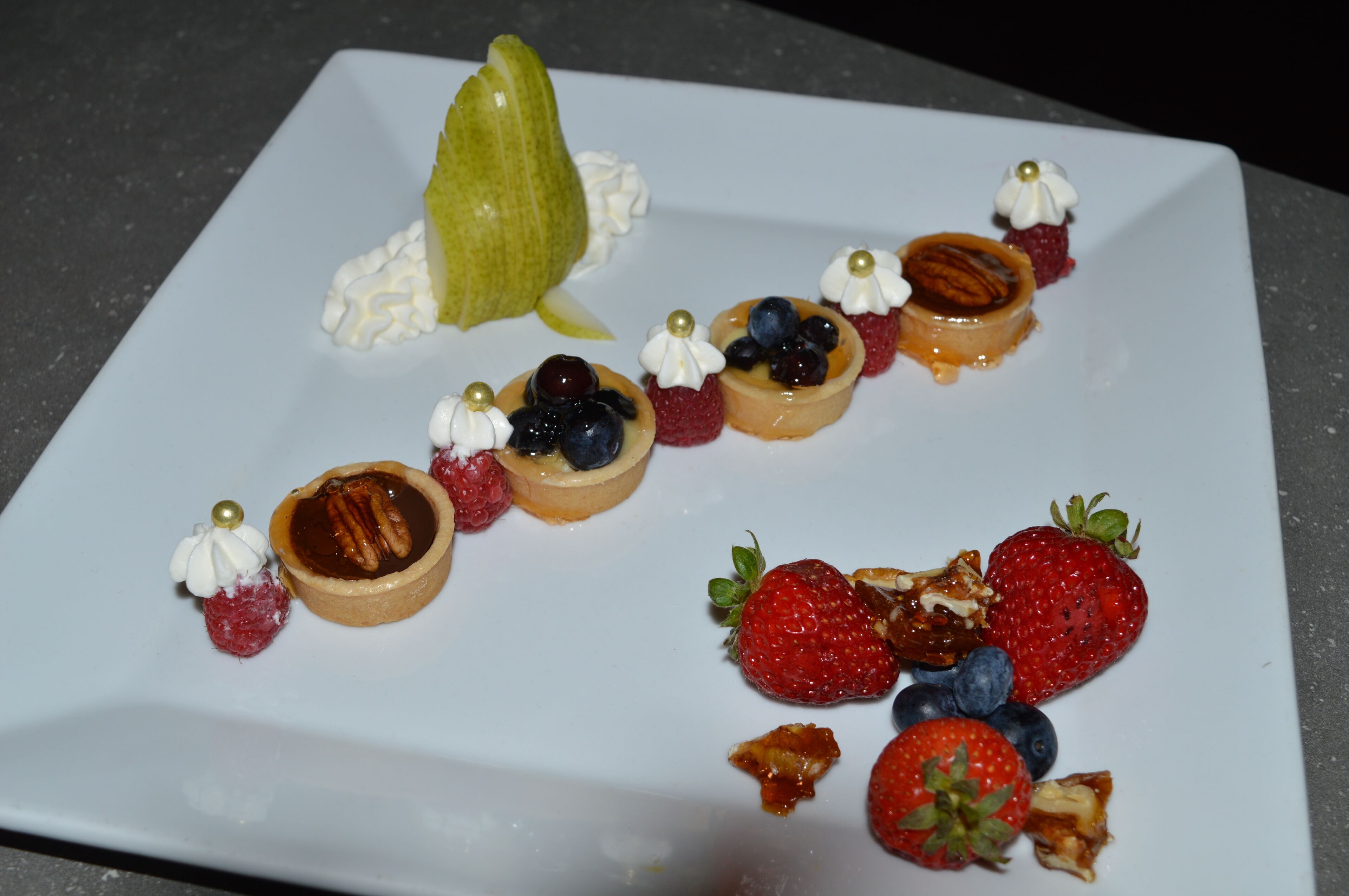 A square white plate with various desserts including tarts, berries, and a pear.