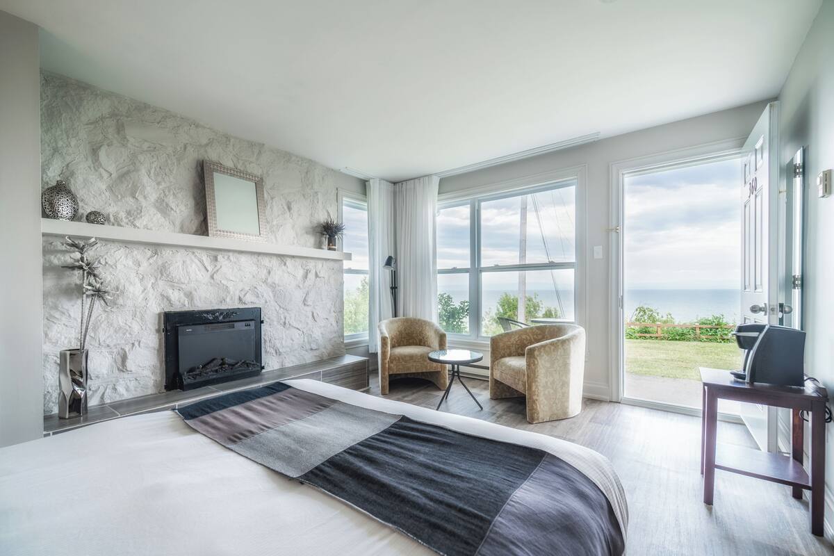 Een moderne slaapkamer met een groot bed, een open haard, twee fauteuils, een bijzettafeltje en uitzicht op de omgeving.