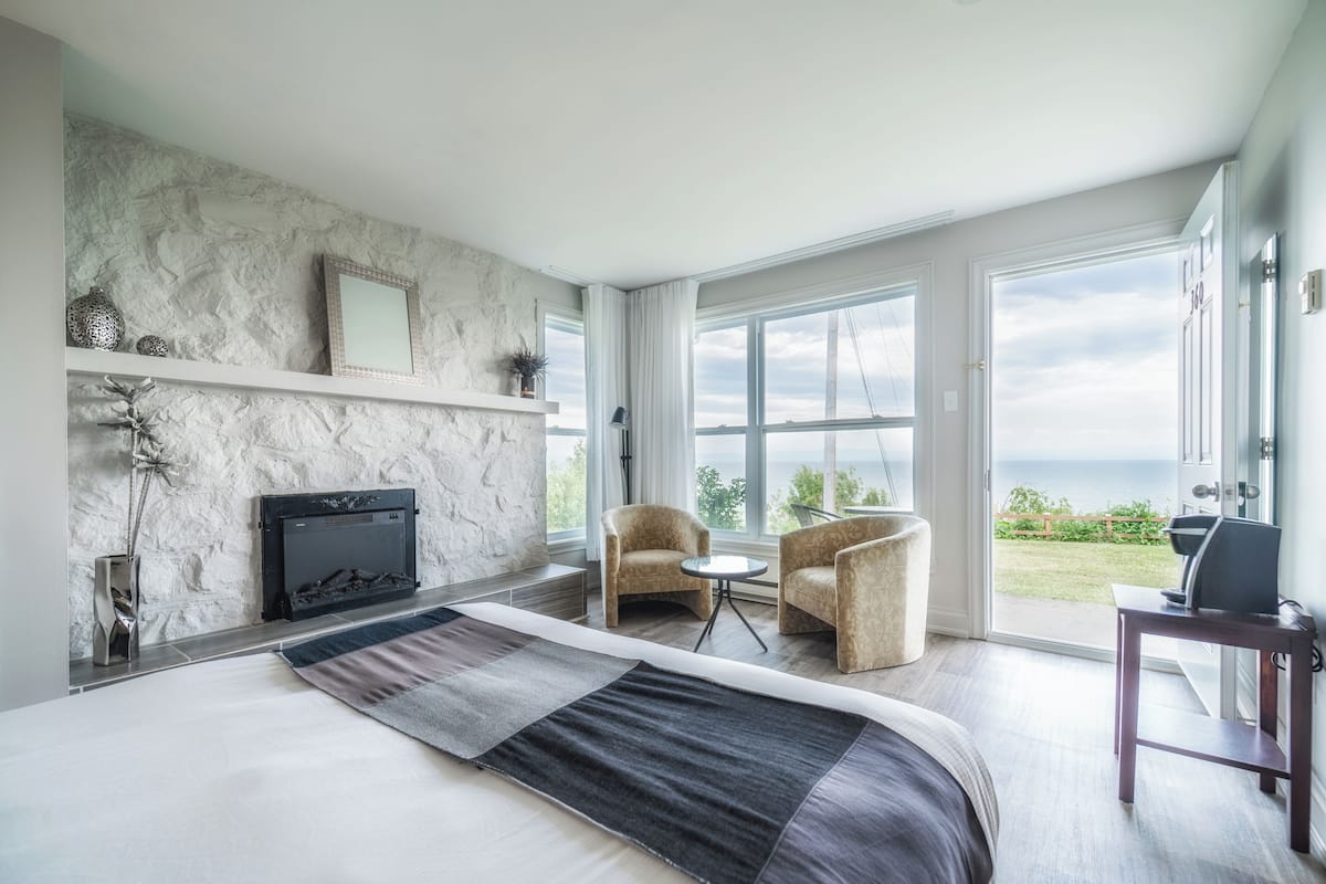 Une chambre moderne avec un grand lit, une cheminée, deux fauteuils, une petite table et une vue sur l’extérieur.