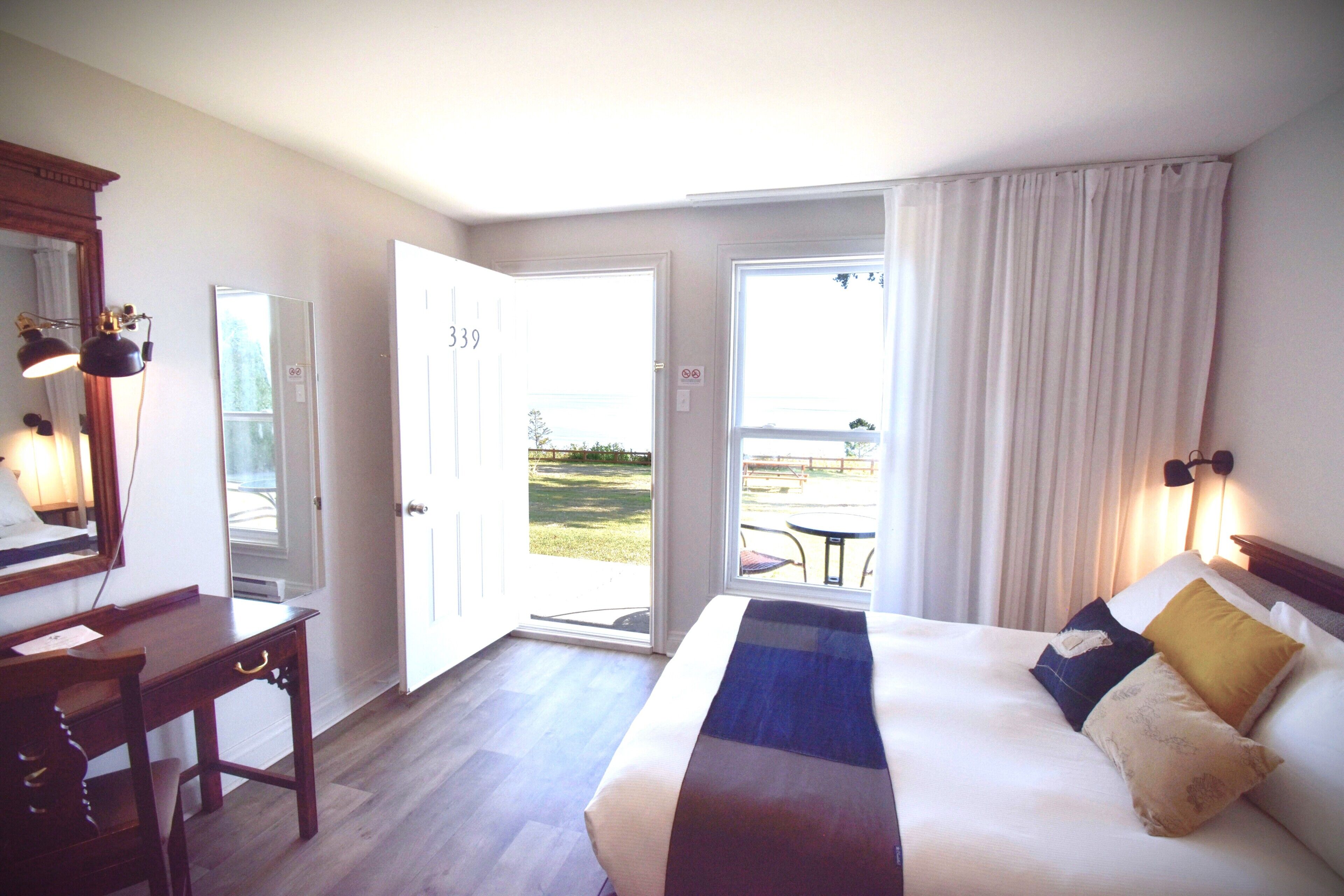 Une chambre d’hôtel comprenant un lit, un bureau, une chaise, un miroir et une porte donnant sur un espace extérieur.