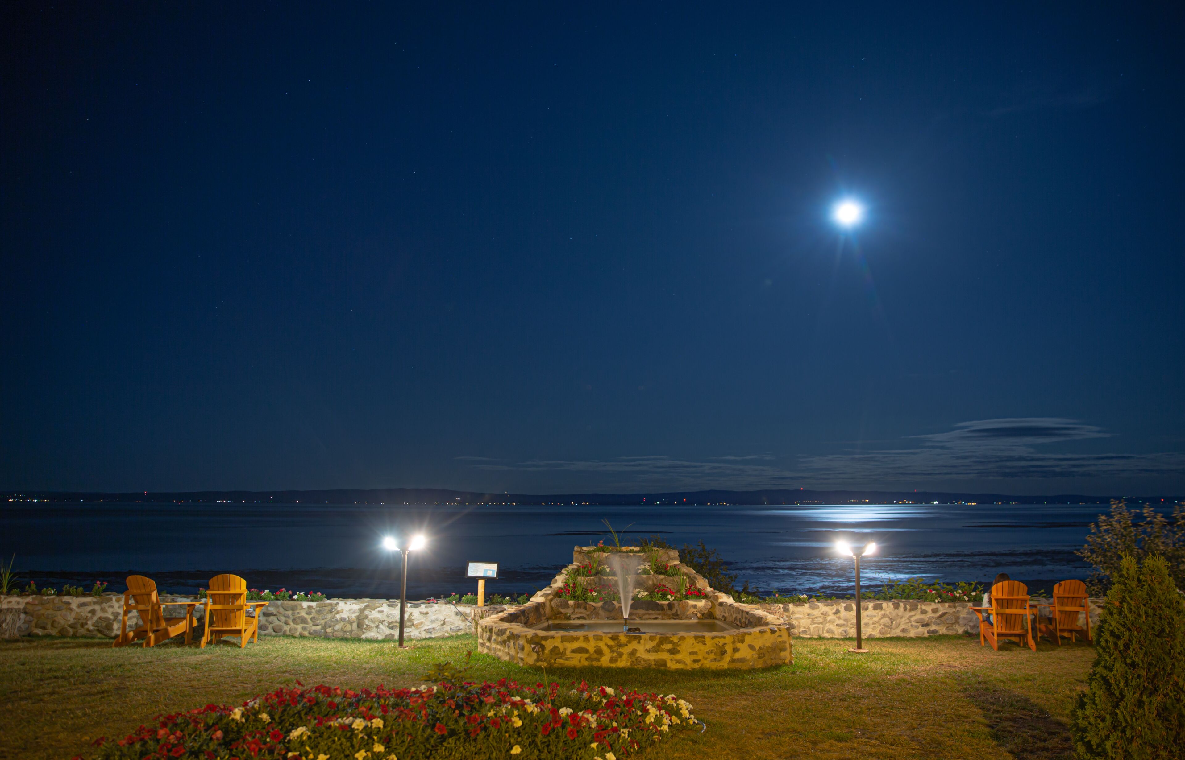 A nighttime outdoor seating area with chairs and a fountain, overlooking a body of water.