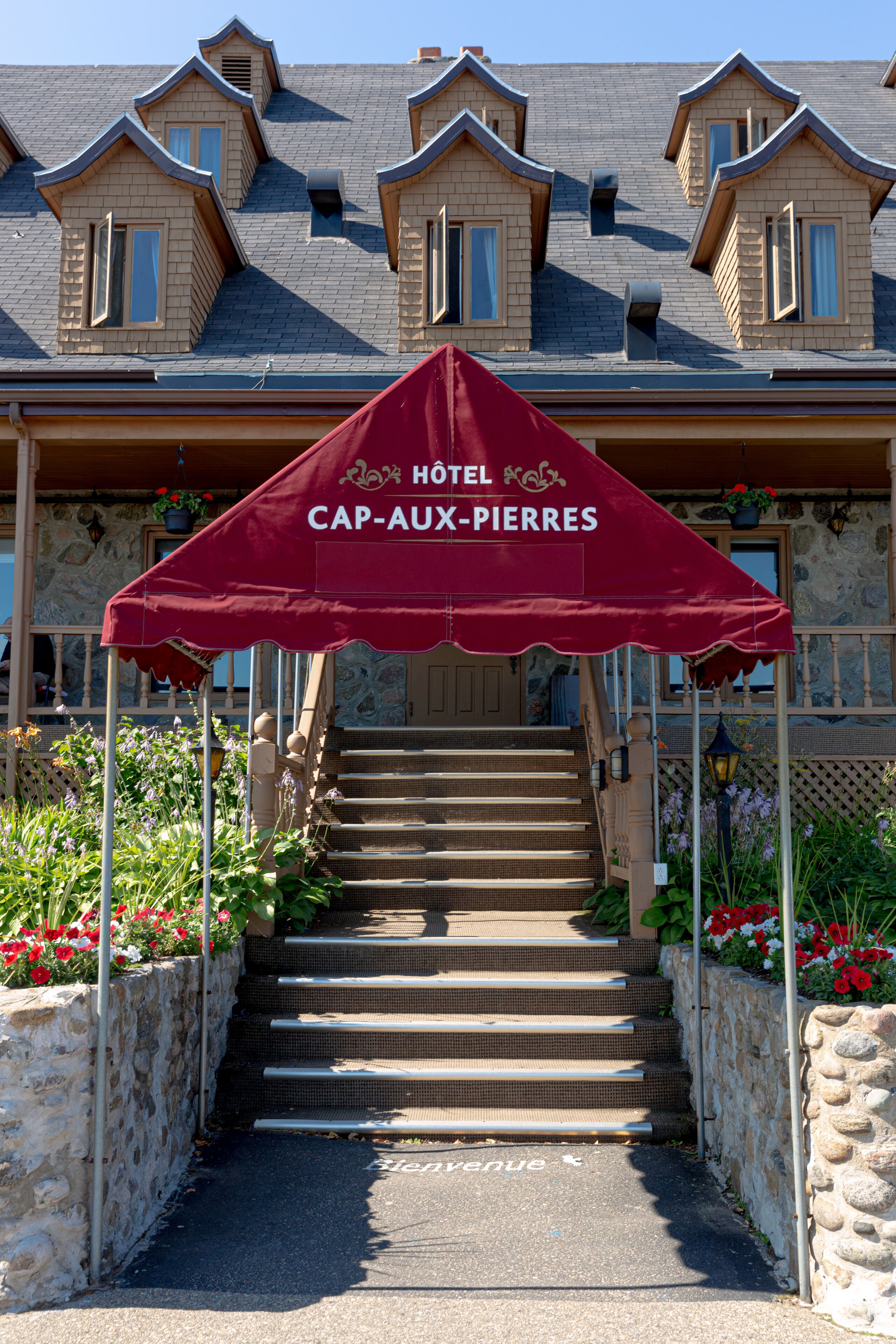 L’entrée d’un hôtel, avec un auvent rouge portant l’inscription « HÔTEL CAP-AUX-PIERRES », des marches en pierre et des parterres de fleurs.