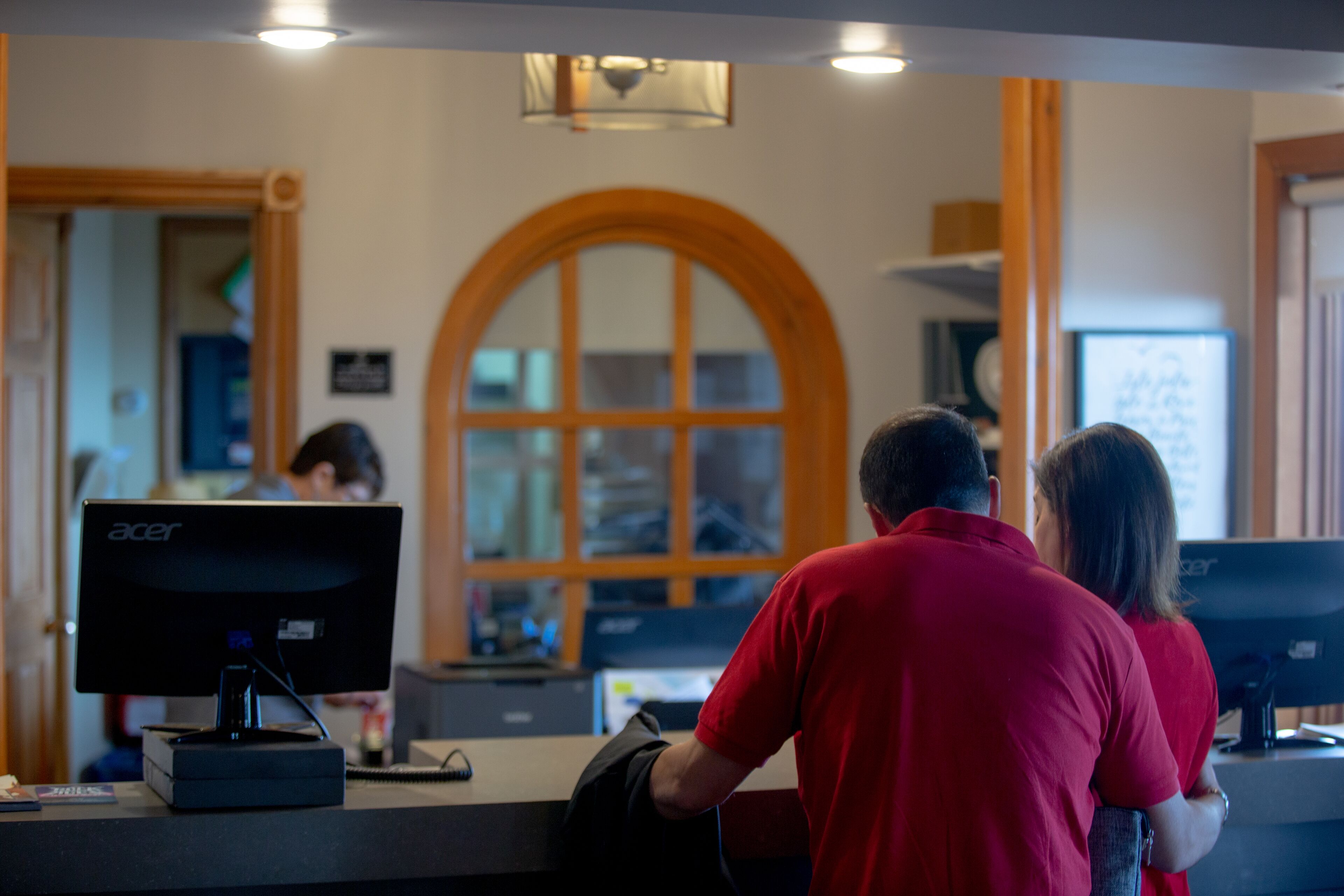 A reception area with a large arched window, a desk with a computer, and a person in a red shirt standing behind the desk.