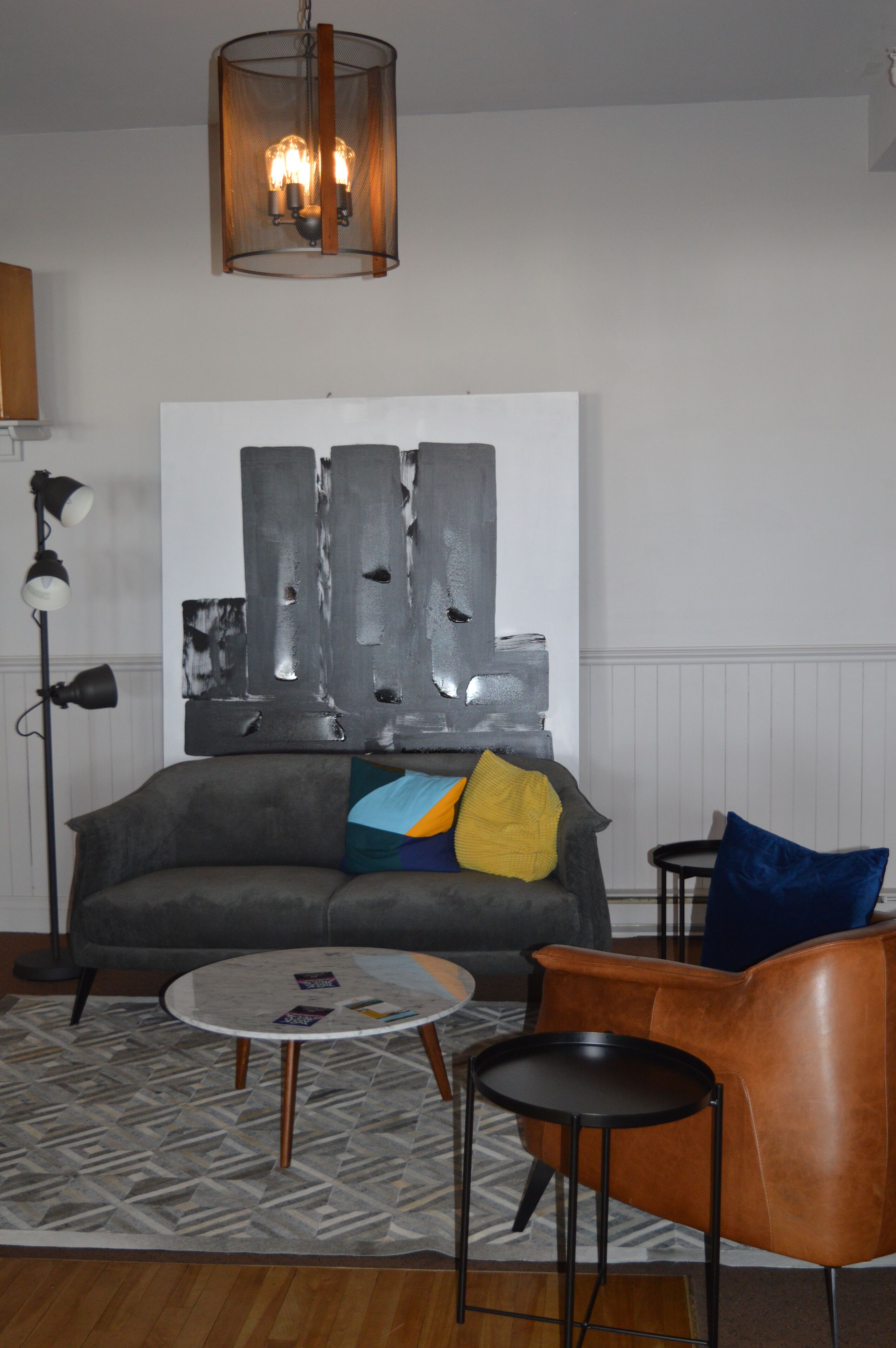 A modern living room with a gray sofa, a round coffee table, and a brown leather armchair. There is a large abstract painting on the wall and a floor lamp.